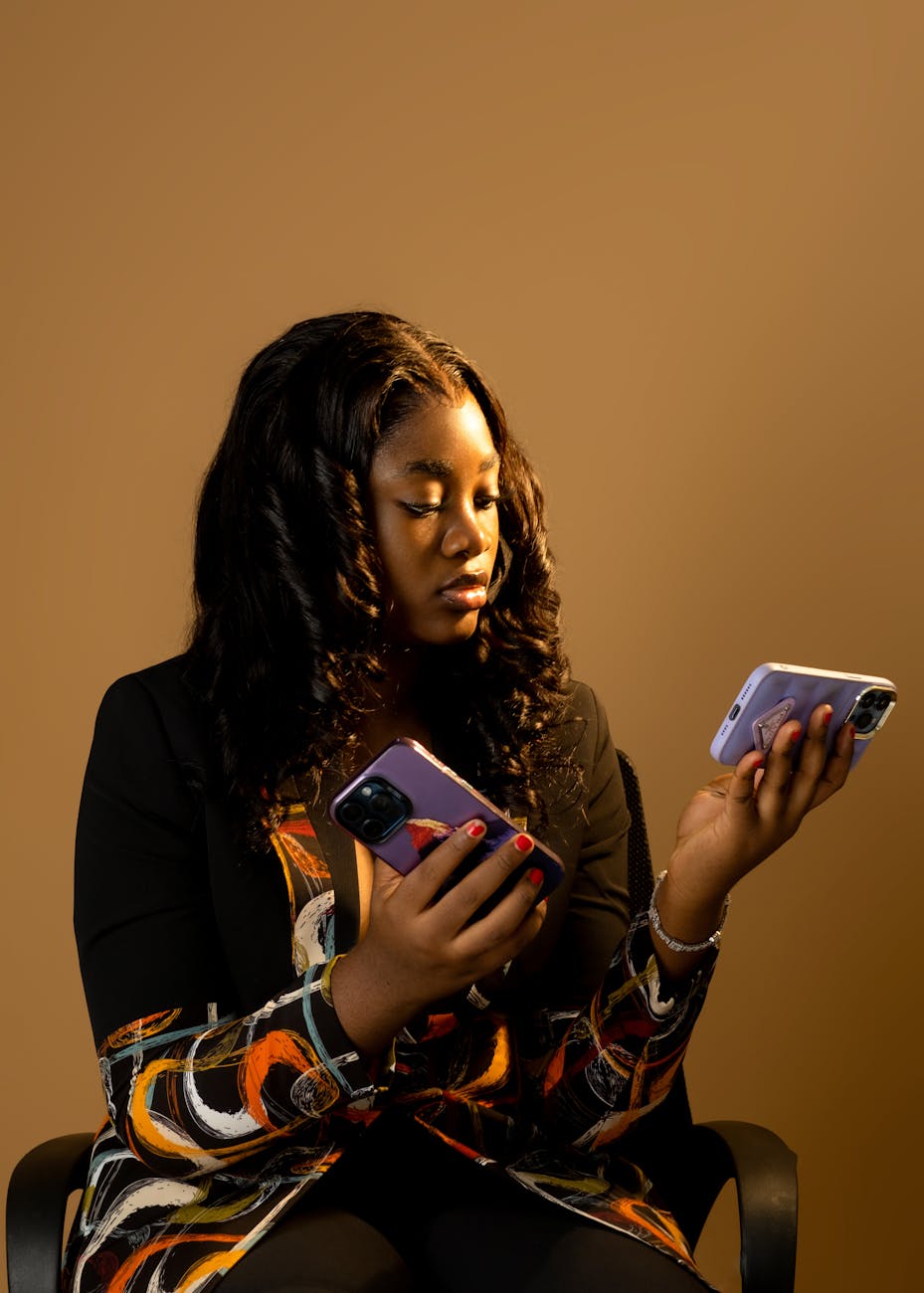 A woman sitting in a chair, holding two smartphones in her hands, with a thoughtful expression against a warm brown background.