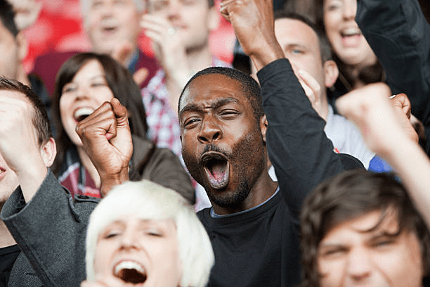 A diverse crowd cheering enthusiastically in a stadium, with a focus on a man in the foreground expressing excitement and encouragement.