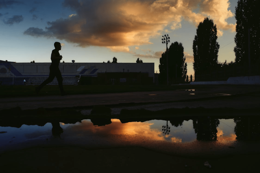 A silhouette of a person jogging along a track, with a sunset casting colorful reflections in a nearby puddle.