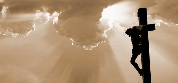 Silhouette of a Jesus Christ on a cross against a dramatic sky with rays of light breaking through the clouds.