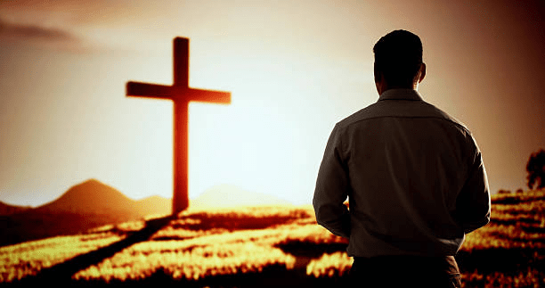 A man stands with his back towards the viewer, gazing at a large cross against a sunset background, symbolizing faith and reflection.