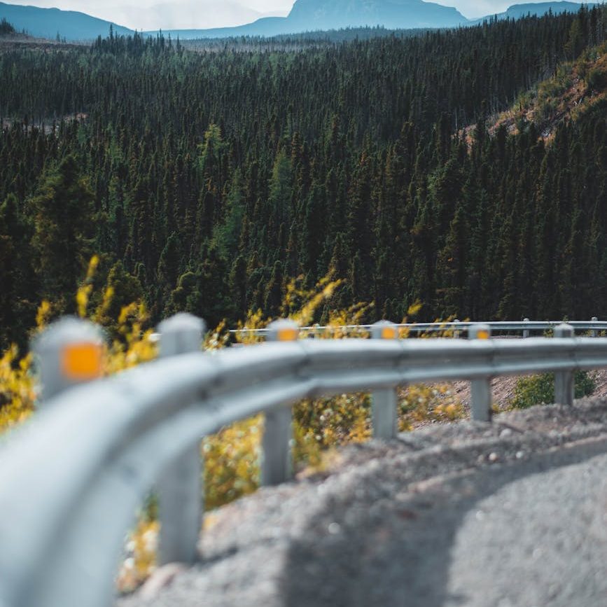 A winding road with a guardrail, surrounded by dense green forests and distant mountains under a cloudy sky.
