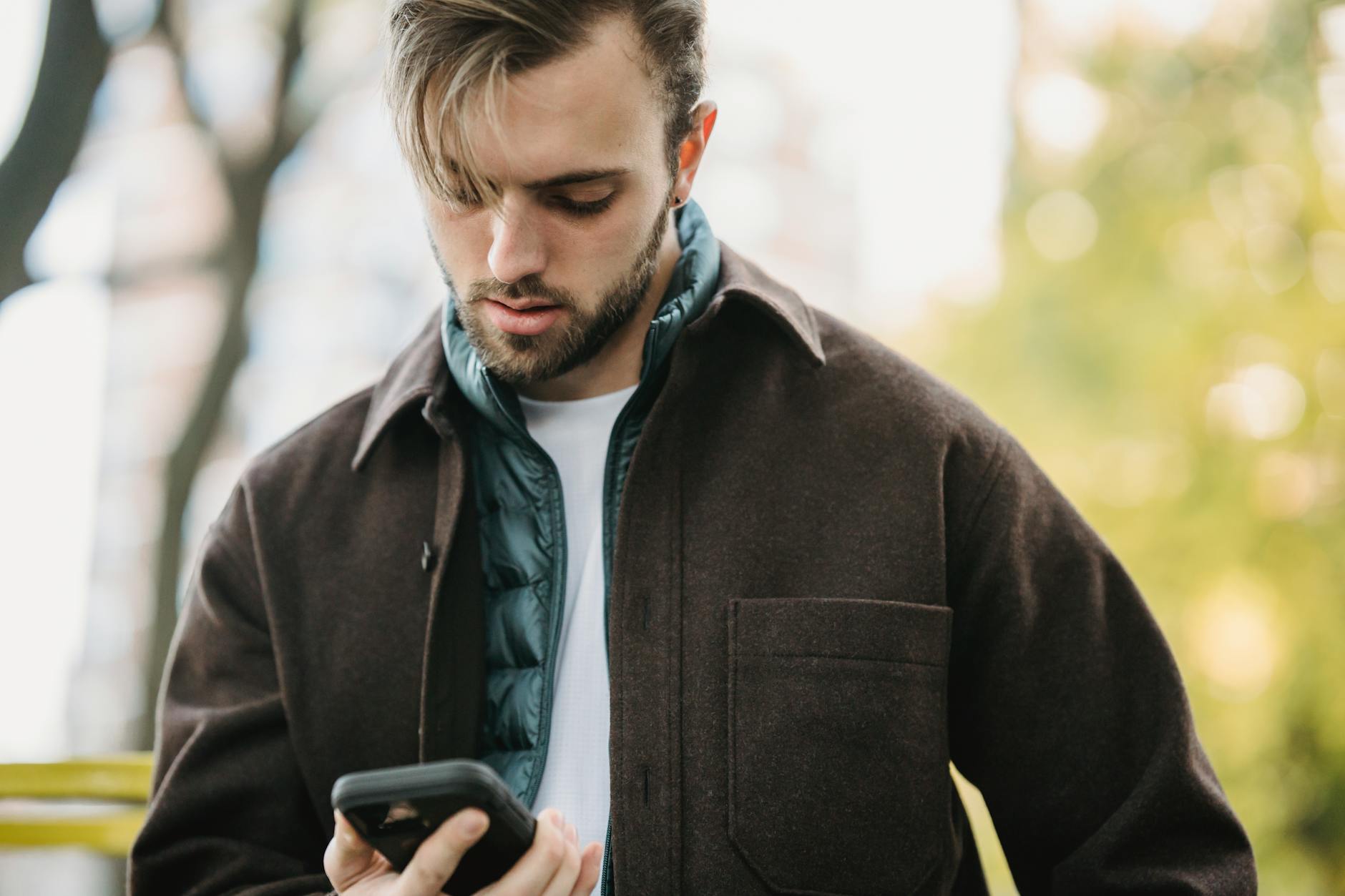 A young man in a brown jacket and a green vest is looking down at his smartphone outdoors, surrounded by blurred greenery in the background.