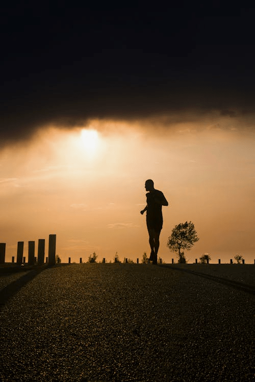 A silhouette of a person jogging on a smooth road during sunset, with the sun partially hidden behind clouds.