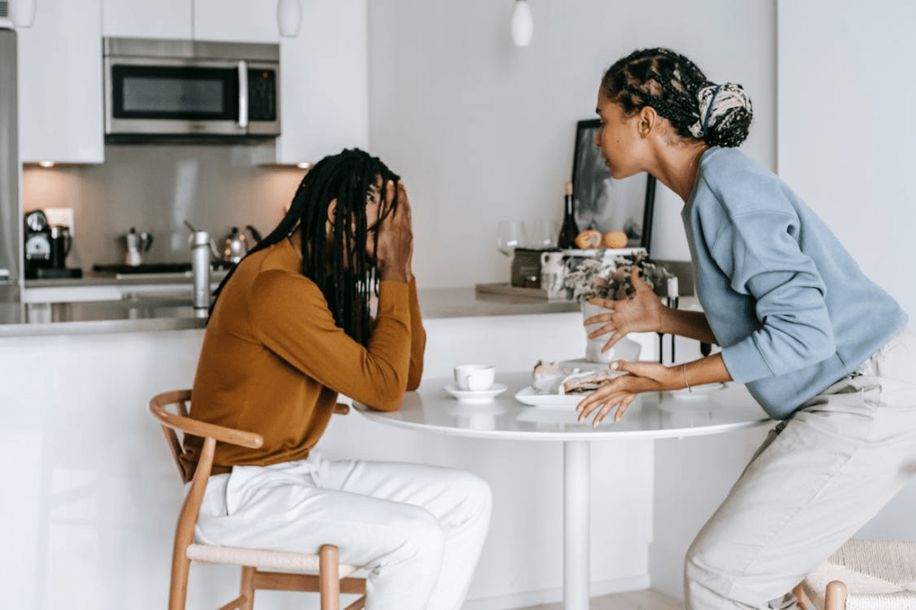A woman with braided hair is expressing herself passionately while sitting at a table across from a man who has his head in his hands, suggesting a tense conversation in a modern kitchen setting.