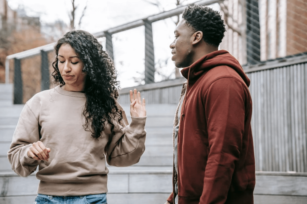 A woman in a beige sweatshirt and a man in a red hoodie stand on outdoor steps, engaged in conversation. The woman appears to be expressing her thoughts while the man listens attentively.