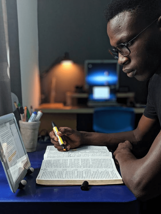 A person studying the Bible while marking using a yellow highlighter and looking intently at the text. A tablet and stationery items are visible in the background, along with a computer.