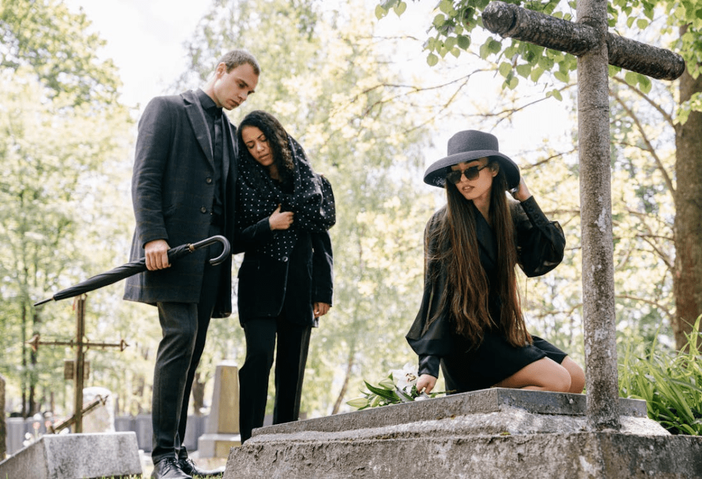 Three individuals dressed in dark attire at a cemetery, with one person seated on a gravestone, holding a flower. The scene features a cross, greenery, and gravestones.