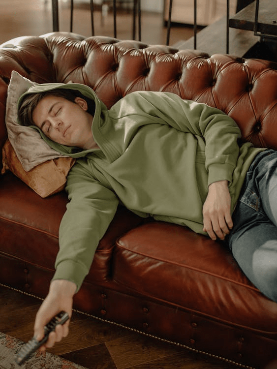 A young man wearing a green hoodie is lying on a brown leather sofa, appearing relaxed or asleep, with a remote control in his hand.