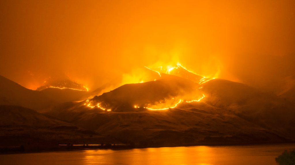 A dramatic landscape featuring mountains engulfed in flames, with an orange sky and smoke. A river is visible in the foreground.