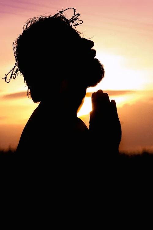 Silhouette of a man with a crown of thorns praying against a sunset background.
