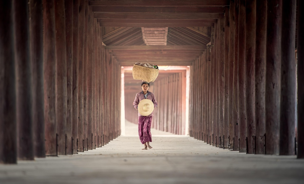 Woman walking barefoot in a long wooden corridor carrying a basket on her head and holding a woven hat