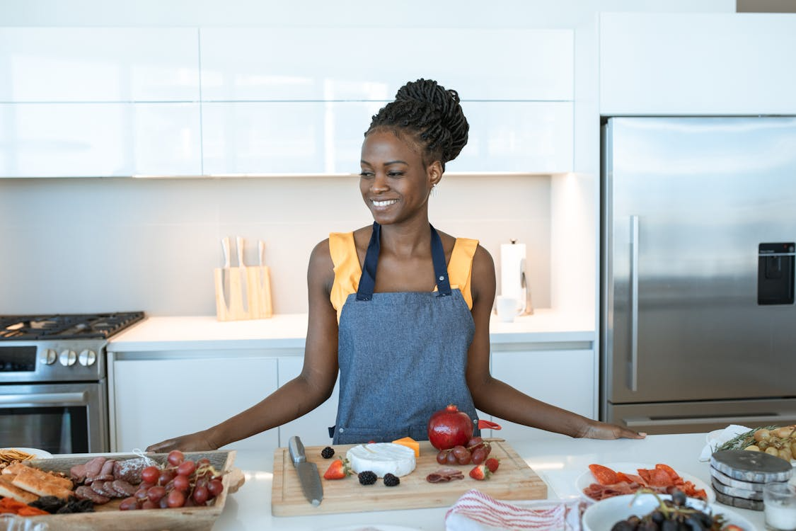 A smiling woman wearing a denim apron stands in a modern kitchen with a cutting board of fruits, cheese, and meats in front of her