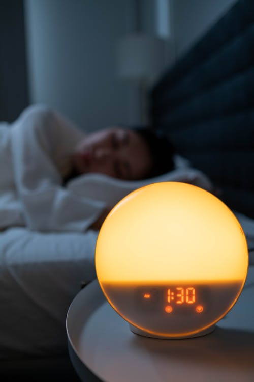 Round bedside alarm clock glowing softly displaying time 1:30 with a person sleeping in the background