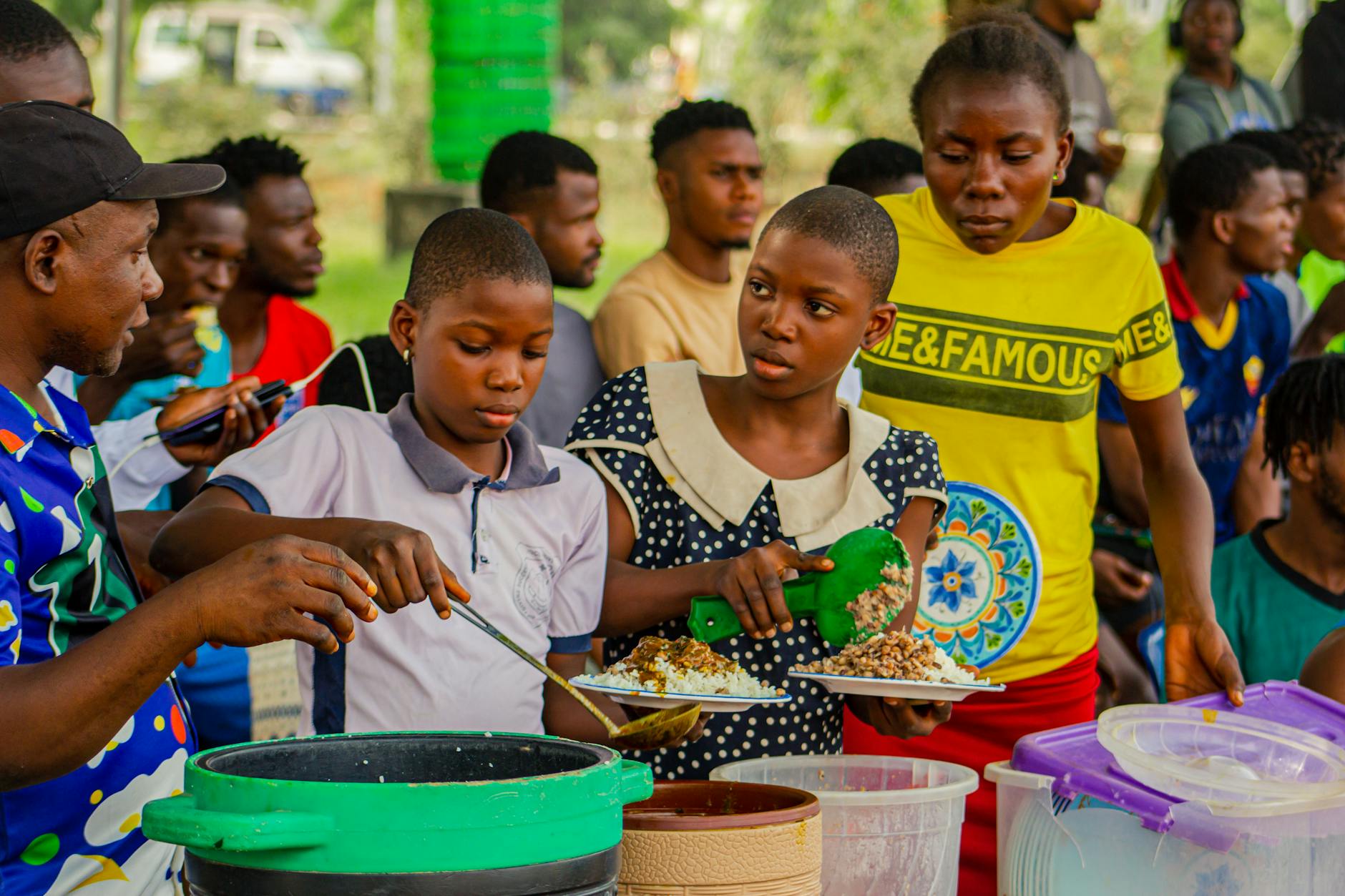 Two children serving food onto plates from large containers at an outdoor gathering