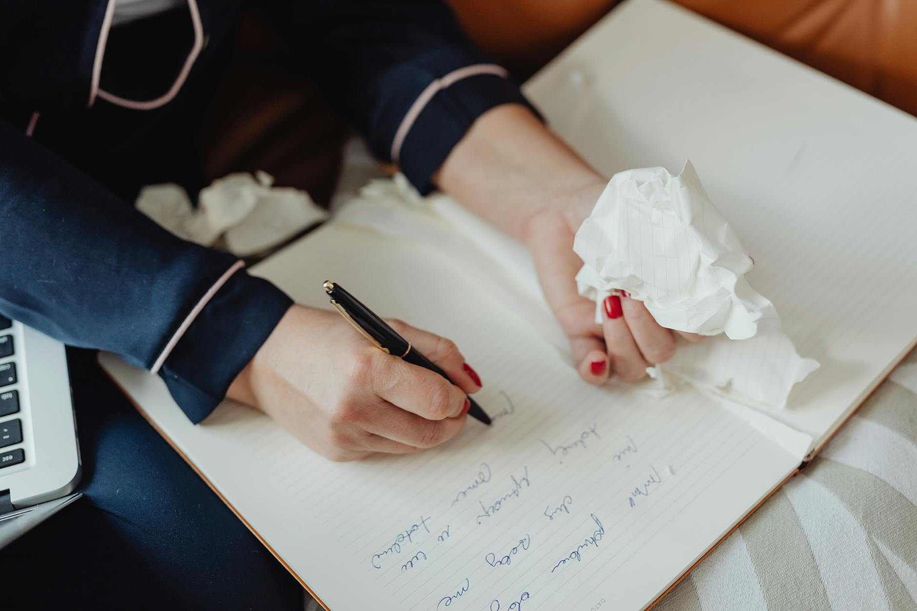 Person writing in a notebook with a pen while holding a crumpled piece of paper