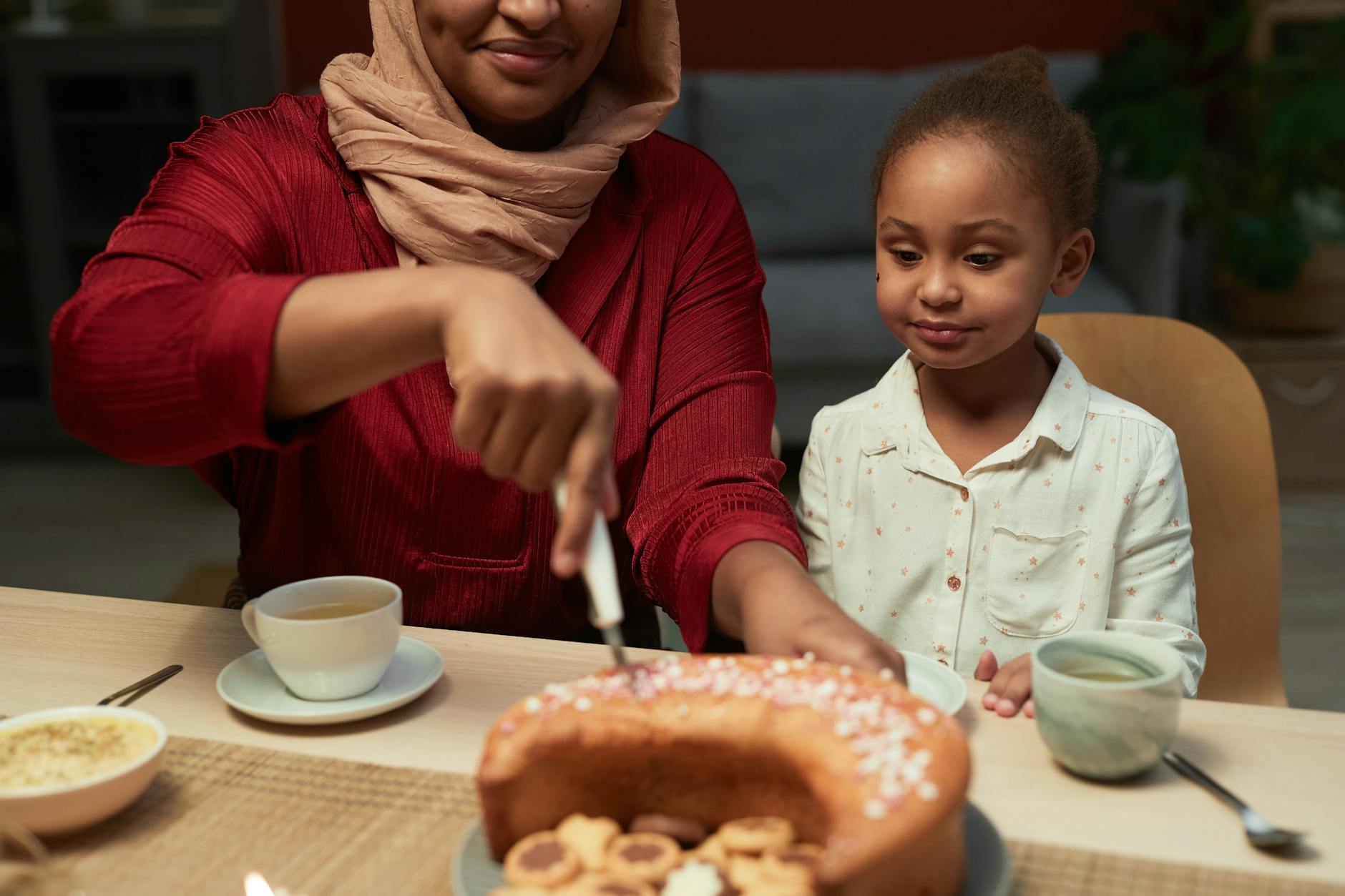Woman wearing a headscarf cutting a cake while a young girl watches at a table with tea cups