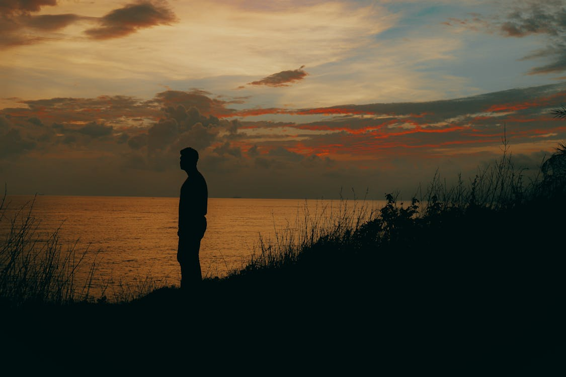 Silhouette of a person standing on a hillside overlooking a calm ocean during a sunset with orange and blue sky