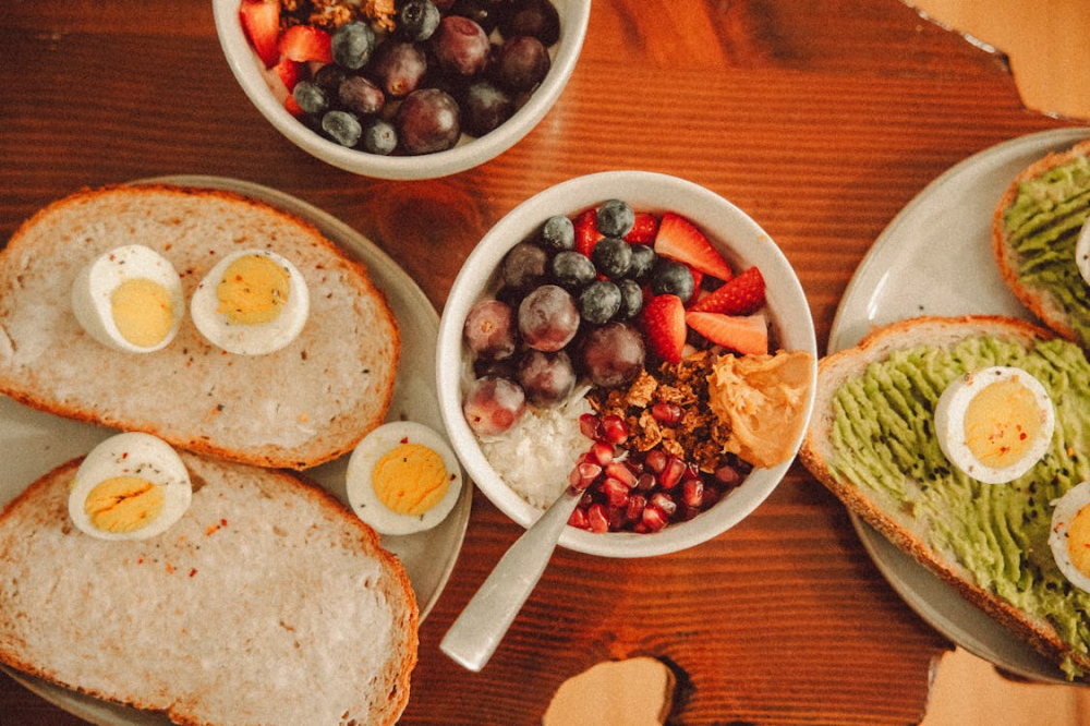 Plates with slices of bread topped with hard-boiled eggs and avocado spread, and bowls containing mixed fruits, granola, peanut butter, and cottage cheese