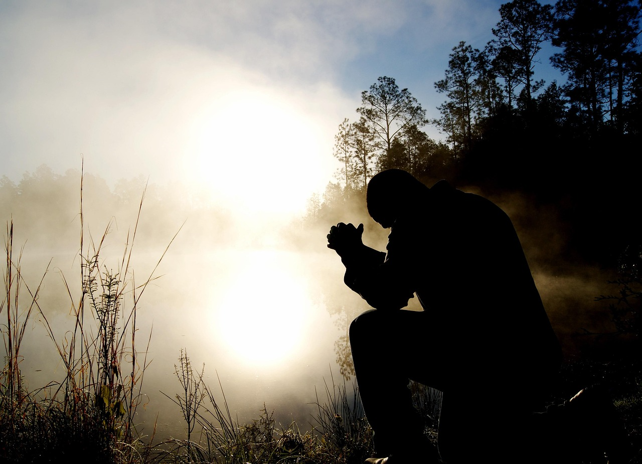 Solitary figure praying by lake Silhouette of a person kneeling and praying by a lake with mist and sunrise