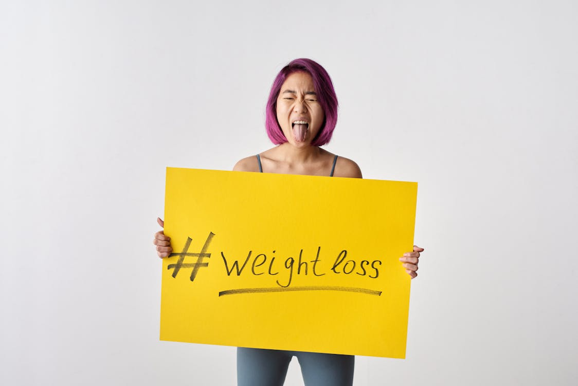 Expression of frustration with weight loss message Person with purple hair sticking out tongue while holding yellow sign reading #Weight loss