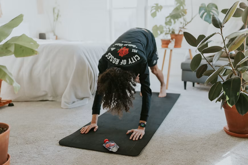 Indoor home exercise session with yoga mat Person in a black City of Roses sweatshirt doing a downward dog pose on a black mat in a bedroom with plants nearby