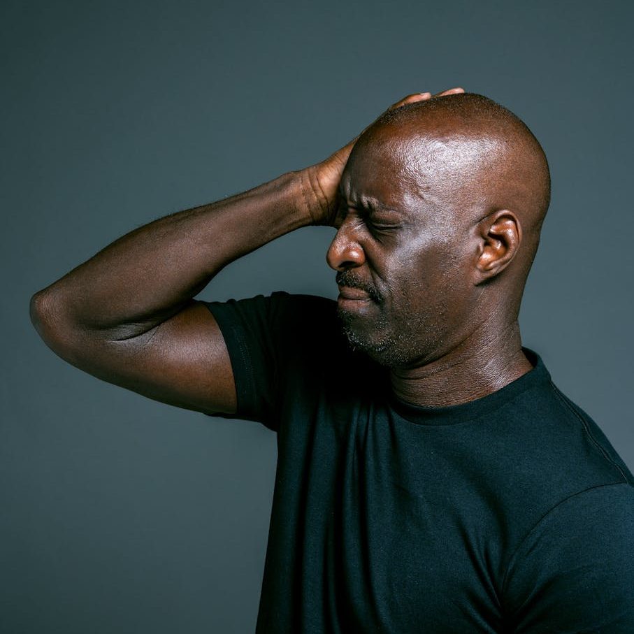 A man in a black shirt with his eyes closed, holding his head with one hand, appearing to be in pain or discomfort