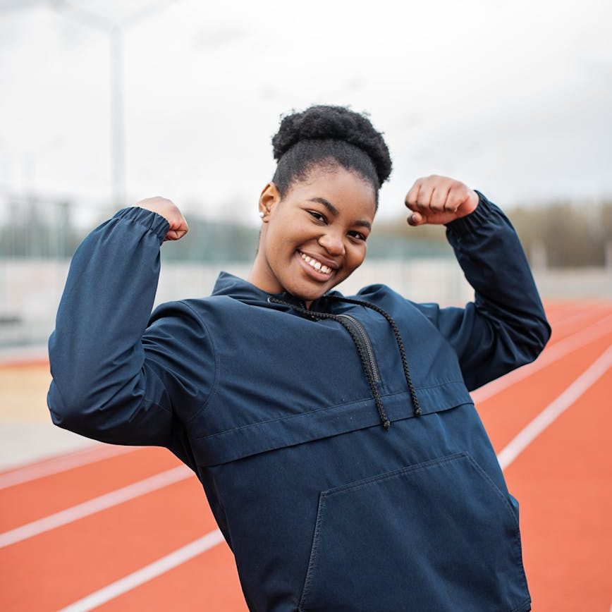 Smiling person in a navy blue jacket flexing their arm muscles on an outdoor running track