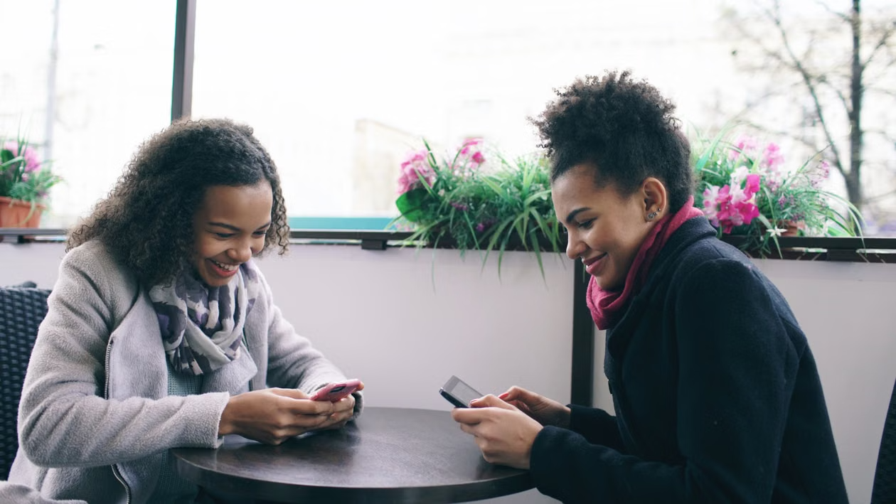 Two women sitting at a round table, smiling and using their smartphones in an indoor setting with plants in the background.