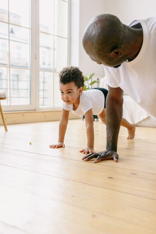 Adult and child in white shirts doing push-ups together on wooden floor near large windows