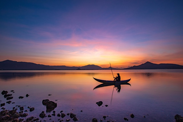 Silhouette of a person paddling a canoe on calm water during a colorful sunset with mountains in the background