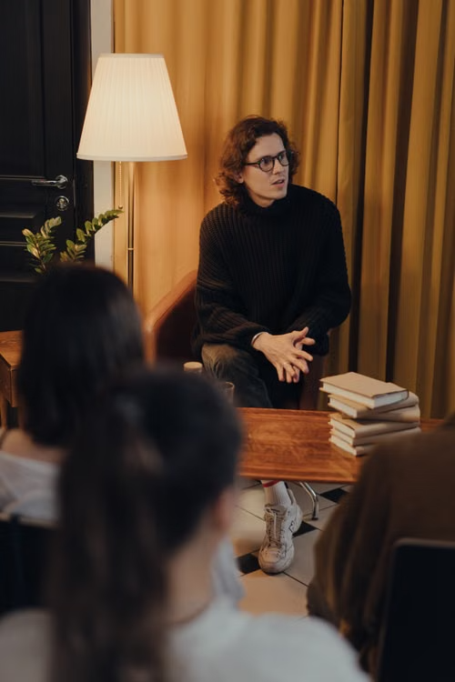 A young man in a black sweater talking to seated people in a cozy room