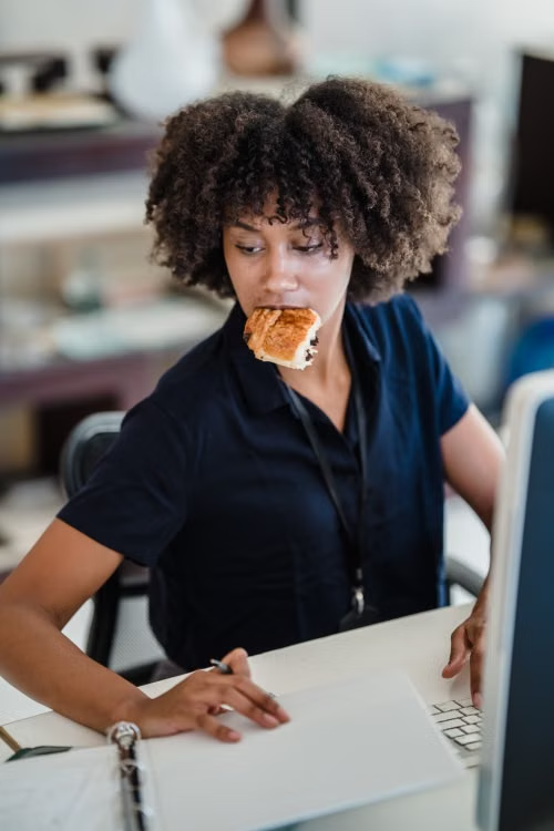 A woman working at a desk with a computer, holding a pen and a pastry in her mouth