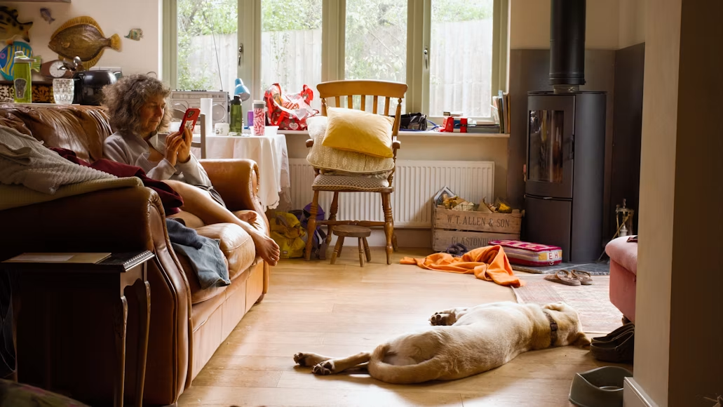 Relaxed home scene with woman and dog A person sitting on a brown leather sofa using a smartphone while a dog lies on the wooden floor in a cozy but cluttered living room with chairs, a wood stove, and various household items