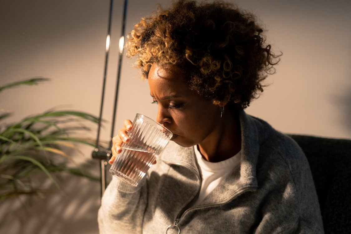 Woman drinking water indoors in soft light Person with curly hair wearing a gray zip-up sweater drinking water from a clear glass indoors