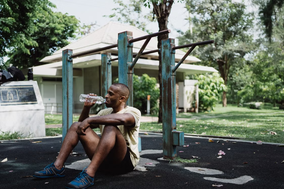 Man resting after outdoor workout A man sitting on the ground drinking water next to outdoor exercise equipment in a park