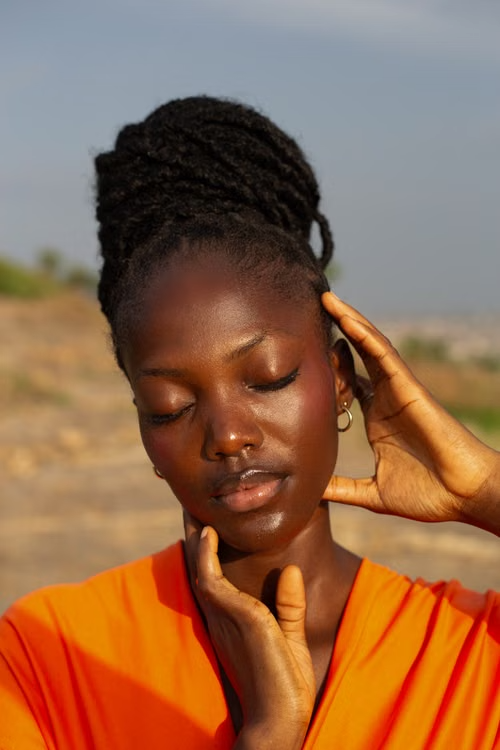 A person with closed eyes touching their face, wearing an orange top outdoors