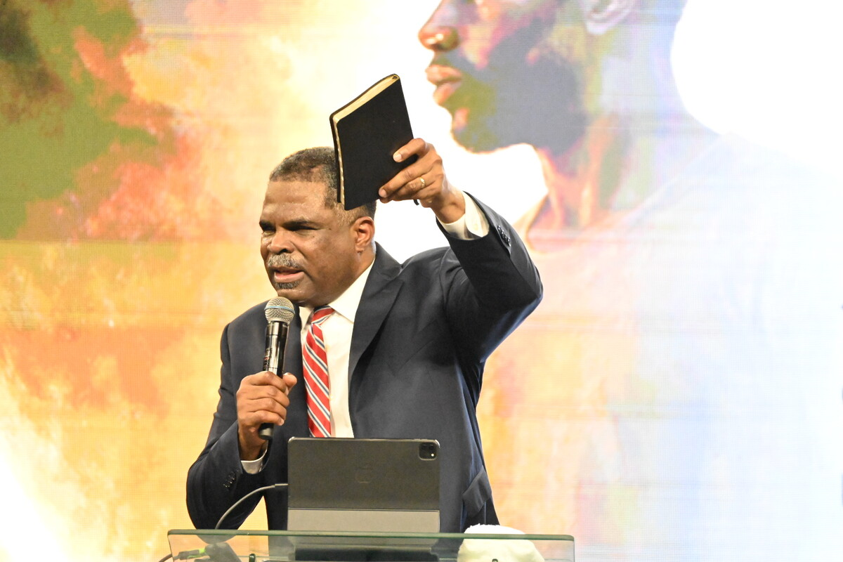 A man in a suit holding a microphone and raising a book while speaking at a podium with a tablet