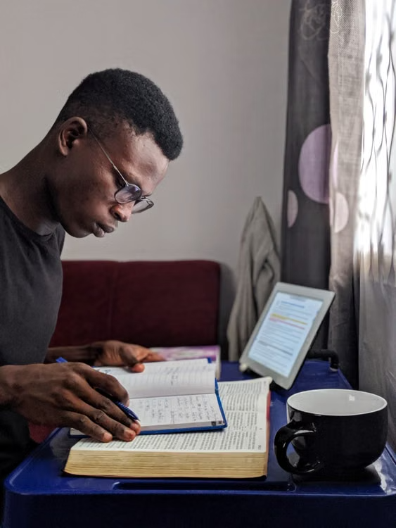 Focused home study with digital reference Student wearing glasses studies from open Bible and a notebook beside a tablet and coffee.