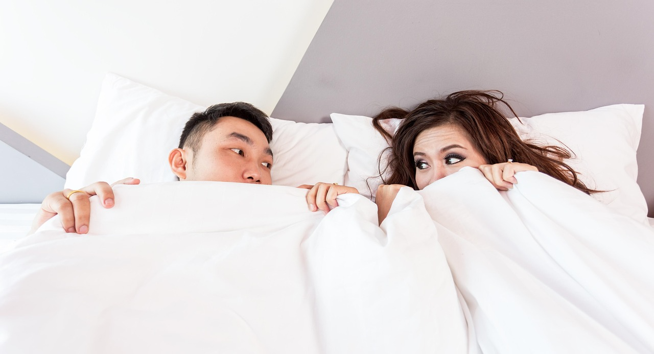 Couple lying in bed, peeking over white blankets and looking at each other