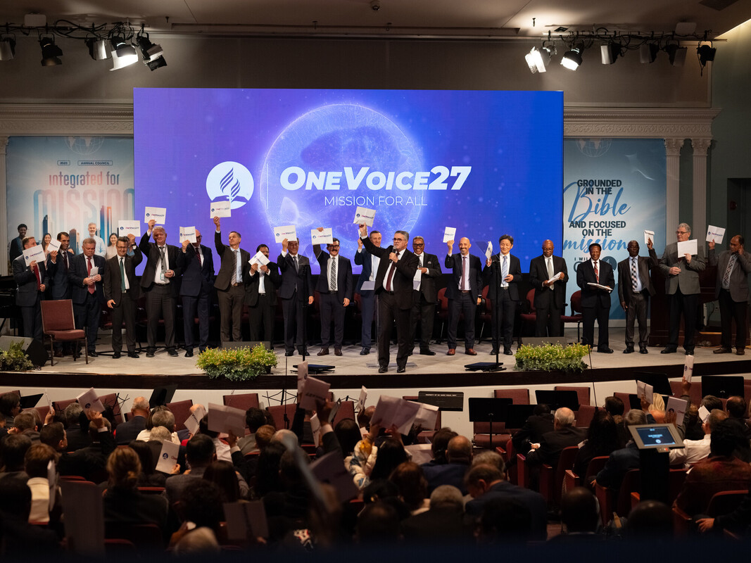 People onstage hold cards beneath a screen reading “OneVoice27: Mission for All.”