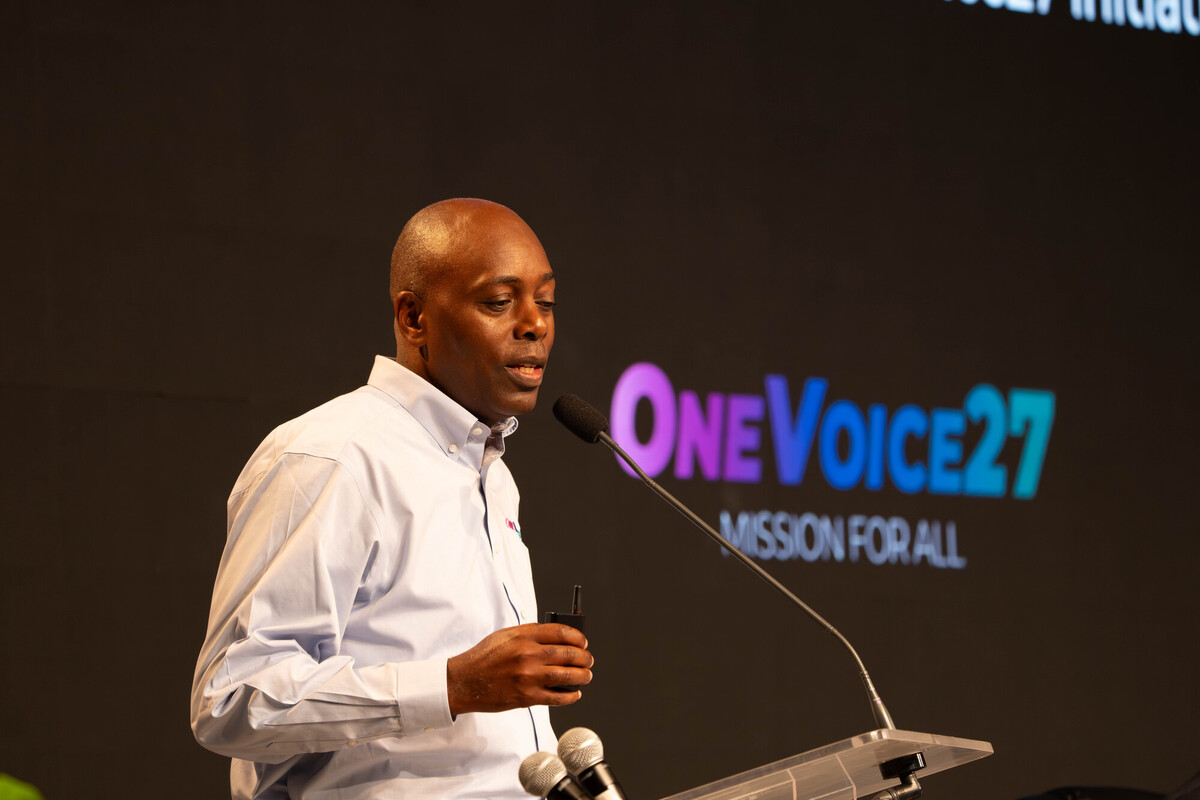 Man speaking at a microphone beside a projected OneVoice27 Mission for All logo