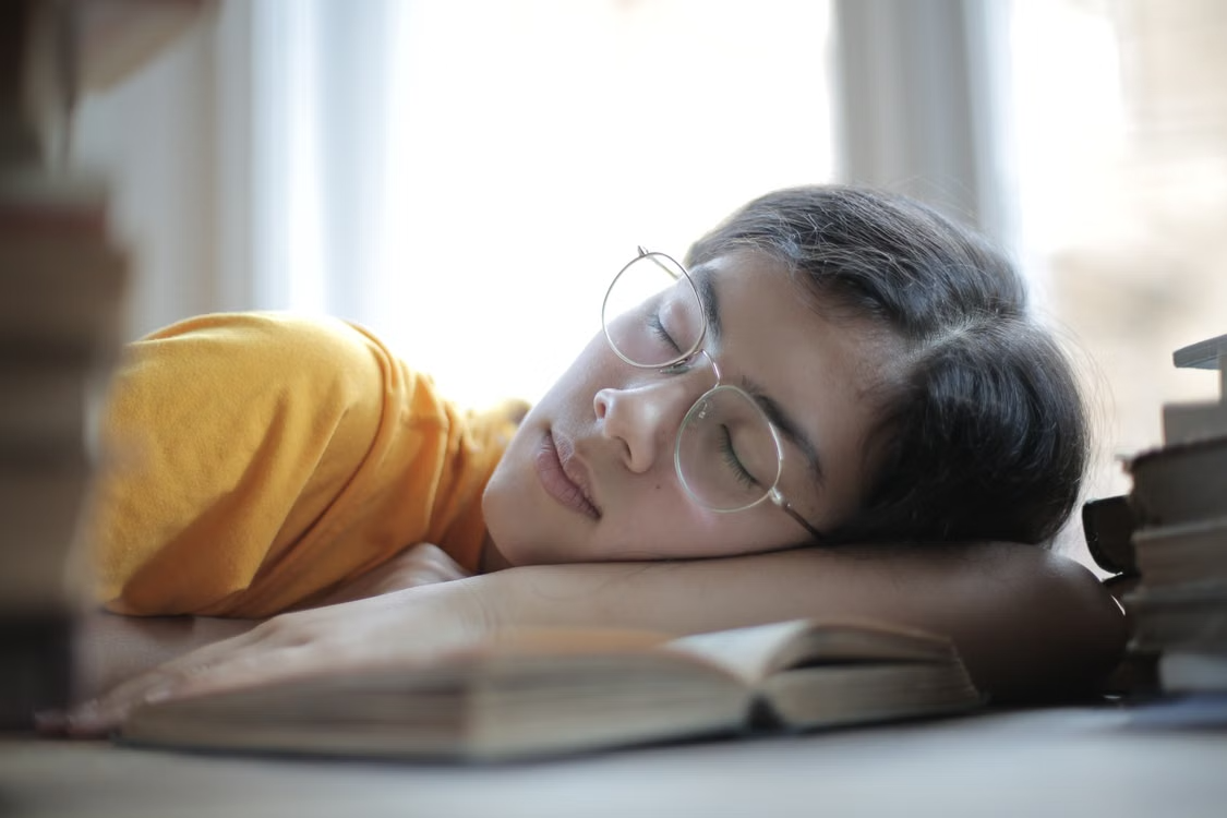 Quiet study break beside open books Person wearing glasses asleep with their head resting on an open book